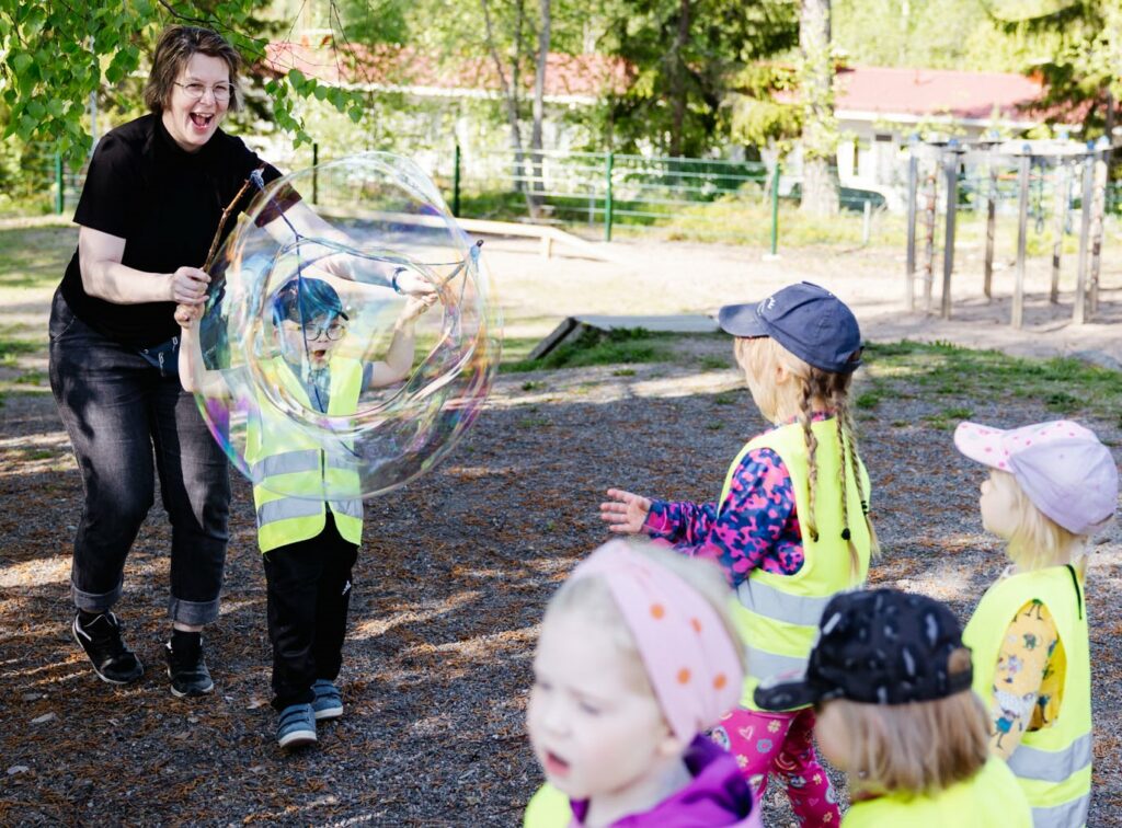 varhaiskasvatuksen lastenhoitaja nauraa lasten kanssa isolle saippuakuplalle